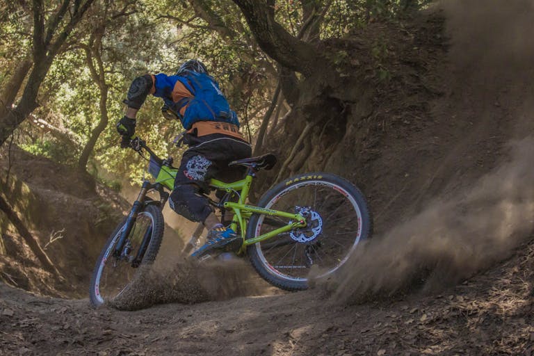 Mountain biker navigates a challenging off-road trail, kicking up dust in a forest setting.